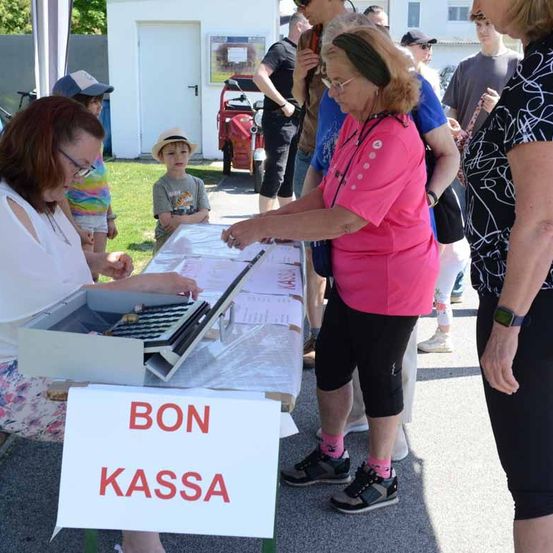 Eine Frau sammelt Geld an einem Tisch mit einem Schild, das 'BON KASSA' steht. Eine Frau in einem weißen Shirt und mit Brille übergibt ihr Geld.