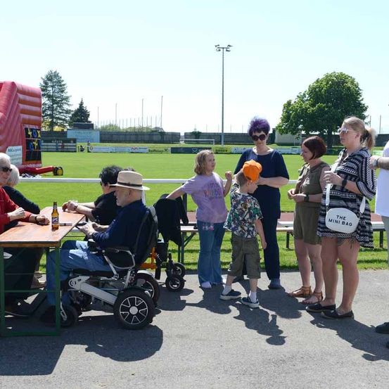 Eine Gruppe von Menschen steht und sitzt im Freien bei einer Veranstaltung, darunter ein Mann im Rollstuhl, eine Frau mit einem Kind und eine Frau mit lila Haaren.