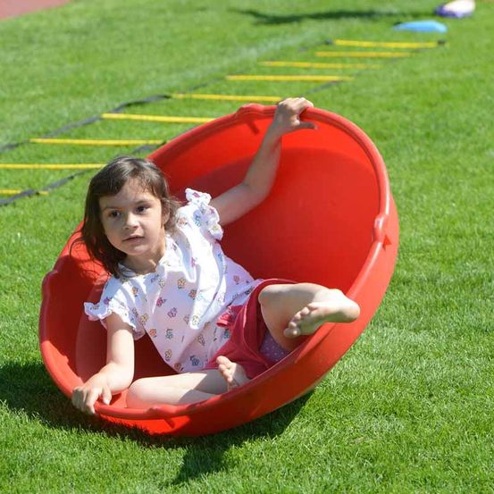 Ein junges Mädchen sitzt in einem roten schüsselförmigen Spielplatzspielzeug auf einem Rasenfeld, mit ausgestreckten Armen.