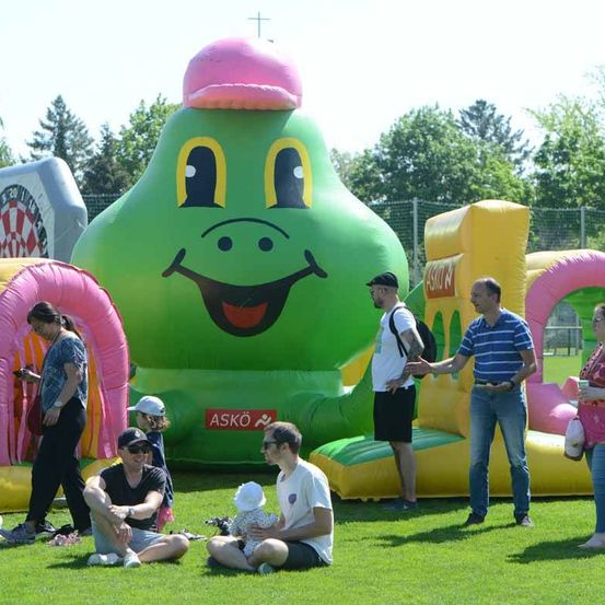 Eine Gruppe von Menschen, die eine Outdoor-Veranstaltung mit einem großen aufblasbaren Frosch, einem Dartboard und einem Spielplatz genießen.