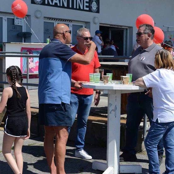 Drei Männer stehen um einen Tisch herum, mit Tassen und einem Mädchen mit geflochtenen Haaren, das in der Nähe geht. Dahinter befindet sich ein Gebäude mit einem Schild, das 'KANTIN' steht.