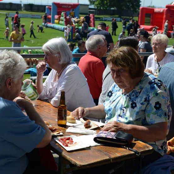 Mehrere ältere Frauen sitzen an einem Tisch im Freien. Sie essen und trinken, mit einer Flasche und einem Becher auf dem Tisch. Im Hintergrund genießen die Menschen den Tag an einem Sprungkasten und auf einer Wiese.