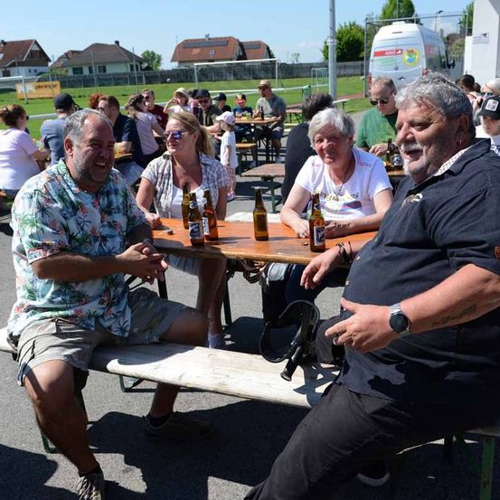 Eine Gruppe von Menschen sitzt an einem Tisch in einem Außenbereich, wobei zwei Männer vorne lächeln und sich unterhalten. Auf dem Tisch stehen Flaschen, und andere sind um sie herum sitzend.