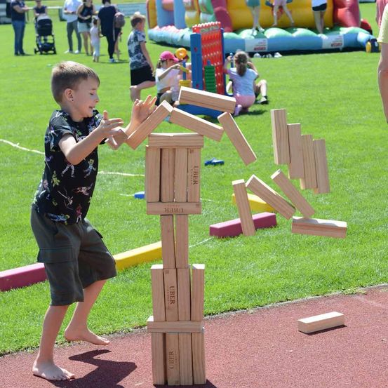Ein Junge versucht, einen Holzturm auf einem Rasenfeld auszubalancieren, mit Menschen und aufblasbaren Spielen im Hintergrund.