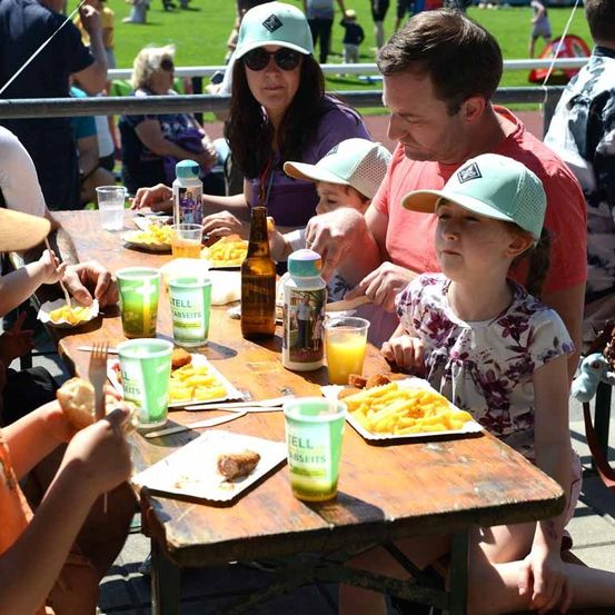 Eine Familie isst bei einer Veranstaltung im Freien. Auf dem Tisch stehen mehrere Teller mit Pommes, Getränken und Essen.