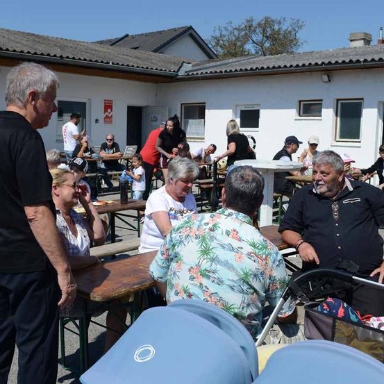 Mehrere Menschen sitzen um Tische herum vor einem Gebäude mit braunem Dach. In der Nähe befinden sich mehrere Kinderwagen.