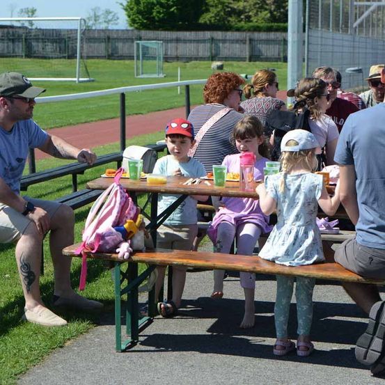 Eine Gruppe von Menschen sitzt an einem Tisch mit Essen und Getränken. Der Mann trägt eine Mütze und Turnschuhe, und die Kinder tragen Mützen. Sie sitzen auf der Bank in einem Außenbereich.