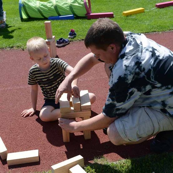 Ein Mann hilft einem Jungen, eine Turm aus Holzklötzen auf einer Laufbahn zu bauen, mit Schuhen und einem Tunnel im Hintergrund.