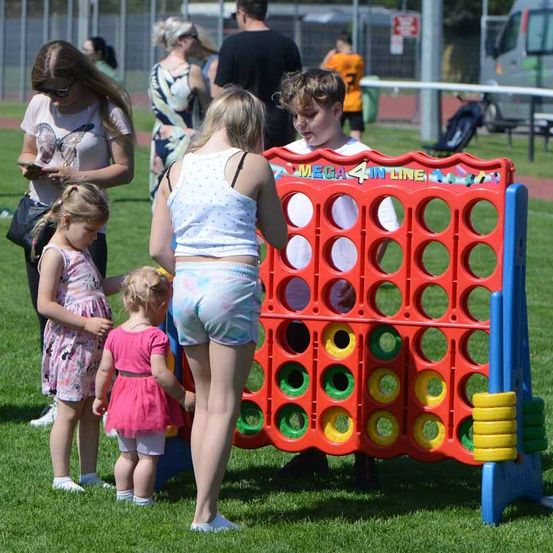 Gruppe von Menschen, die an einem sonnigen Tag Connect Four spielen. Ein Junge und zwei Mädchen spielen, während andere zuschauen.