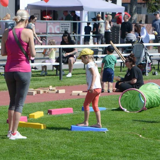 Eine Gruppe von Menschen versammelt sich für einen Familienspaßtag, mit Kindern, die auf einem Rasenfeld spielen, einer Frau mit einem Kinderwagen und anderen, die im Hintergrund zusehen.