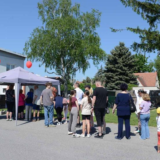 Eine Gruppe von Menschen steht in einer Schlange unter einem lilafarbenen Zelt an einem sonnigen Tag. Im Hintergrund befinden sich Bäume und Häuser.