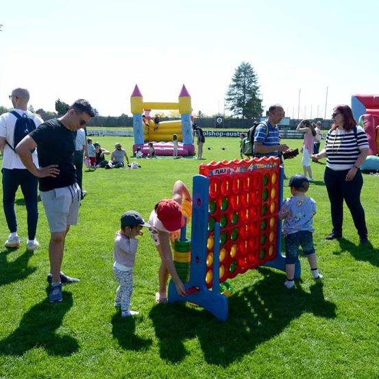 Menschen spielen auf einem Rasen mit aufblasbaren Spielzeugen im Hintergrund. Ein Kind versucht, ein Brettspiel zu spielen, während ein Erwachsener hilft.