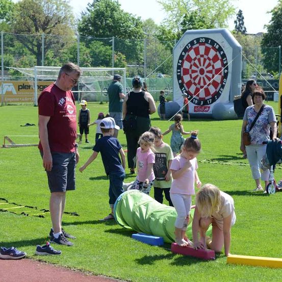Eine Gruppe von Kindern und Erwachsenen spielt auf einem Feld. Ein Mann und eine Frau beobachten die Kinder. Ein Kind spielt auf dem Dartboard und das andere auf dem Boden.