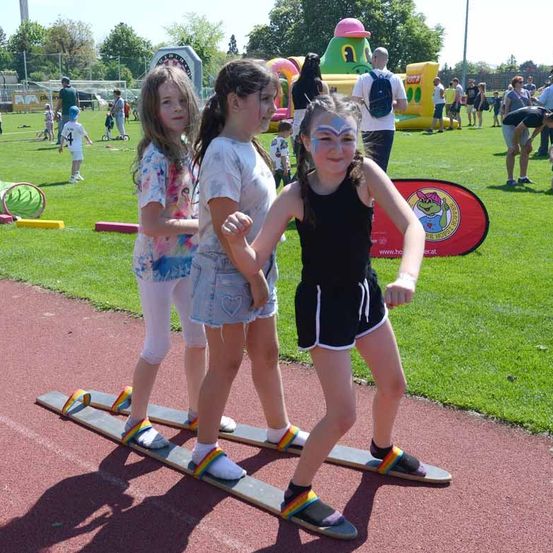 Drei Mädchen laufen auf Holzbrettern auf einer Laufbahn, mit Menschen und aufblasbaren Spielgeräten im Hintergrund.