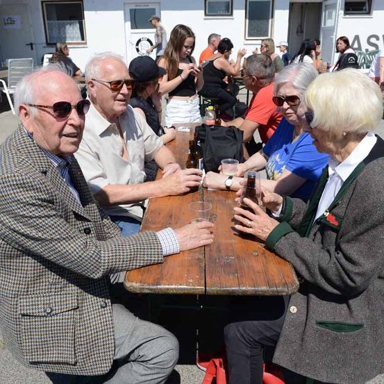Eine Gruppe älterer Erwachsener sitzt in einem Außenbereich an einem Holztisch, trägt Sonnenbrillen und Jacken, mit einem weißen Gebäude und Menschen im Hintergrund.
