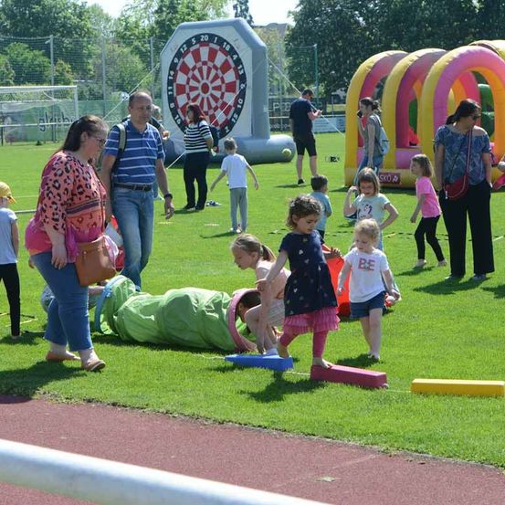 Erwachsene und Kinder spielen auf einem Feld mit verschiedenen aufblasbaren Spielzeugen, einschließlich eines großen Dartspiels, während andere im Hintergrund zuschauen.