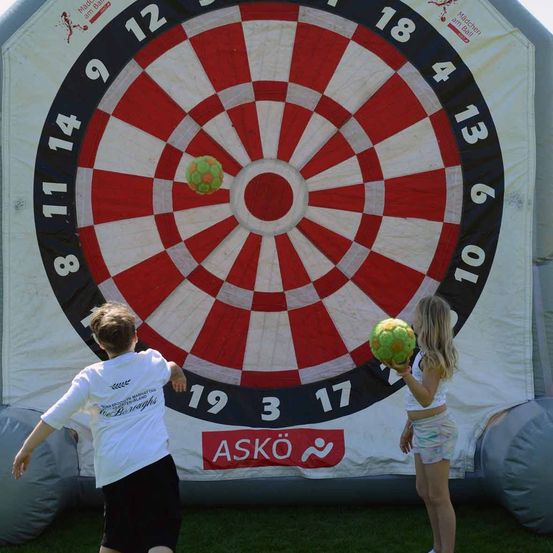 Zwei Kinder stehen vor einem riesigen aufblasbaren Dartboard, jedes hält einen grünen Ball. Der Junge wirft seinen Ball auf das Dartboard, während das Mädchen ihren Ball zum Werfen vorbereitet.