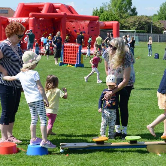 Mehrere Erwachsene und Kinder spielen auf einem großen Rasenfeld. Einige spielen auf einem Wippgerät und andere gehen zu einem Hüpfburg. Einige Kinder rennen.