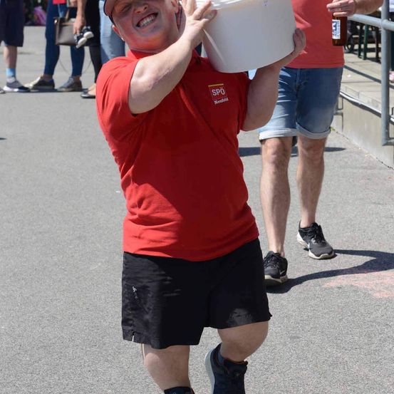 Ein Mann mit einem roten T-Shirt mit dem Aufdruck 'SPO' trägt einen weißen Eimer mit ausgestreckten Armen. Er lächelt und schaut auf etwas. Hinter ihm ist eine verschwommene Ansicht von Menschen, die auf der Straße gehen.