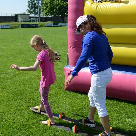 Eine Frau hilft einem Mädchen, auf einem Balancebrett über ein Grasfeld zu gehen, wobei ein buntes aufblasbares Hüpfhaus im Hintergrund steht.