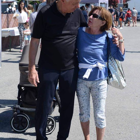 Ein älteres Paar geht draußen spazieren. Der Mann trägt ein schwarzes Shirt und eine Hose, die Frau ein blaues Oberteil, eine Hose und eine Sonnenbrille. Hinter ihnen steht ein Kinderwagen und eine Frau mit einem Baby.