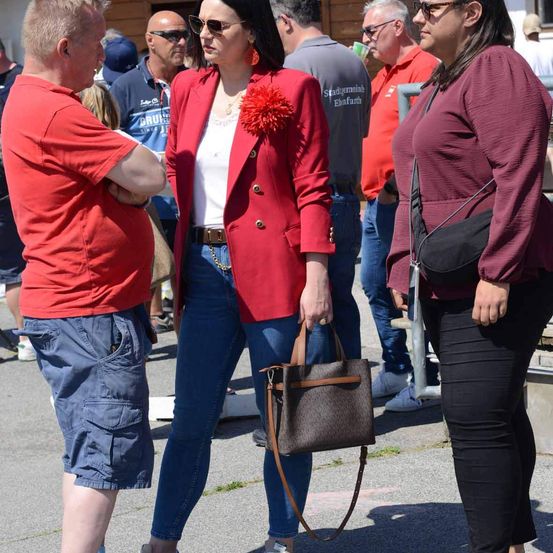 Eine Frau in einer roten Jacke mit einer Blume darauf steht neben einer anderen Frau in einem weinroten Shirt. Sie tragen beide Handtaschen und scheinen mit einem Mann in einem roten Shirt zu sprechen.