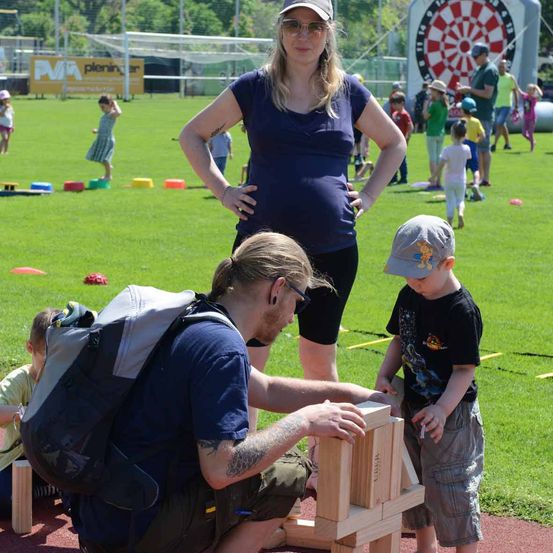 Eine Frau mit Babybauch beobachtet einen Mann und einen Jungen, die auf einem Feld eine Holzstruktur bauen, während andere Kinder im Hintergrund spielen.