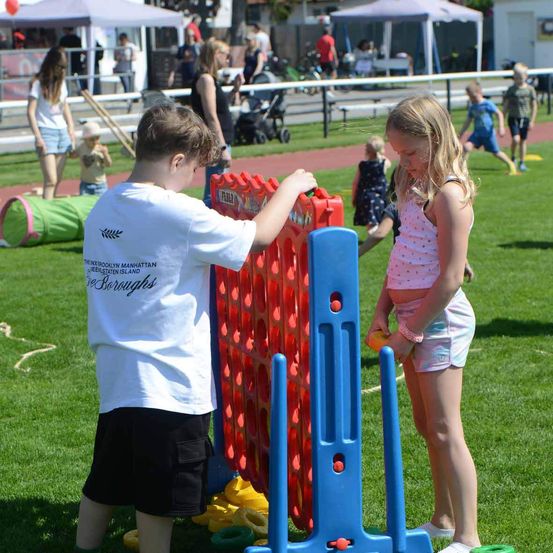 Zwei Kinder spielen auf einem Rasenfeld eine Partie Connect Four. Im Hintergrund sind andere Kinder und Erwachsene zu sehen.