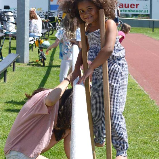 Ein junges Mädchen in einem karierten Overall hält sich an einem weißen Pfosten fest, während eine andere Person ihr hilft. Im Hintergrund sind Fahrräder, eine Bank und eine sitzende Frau zu sehen.