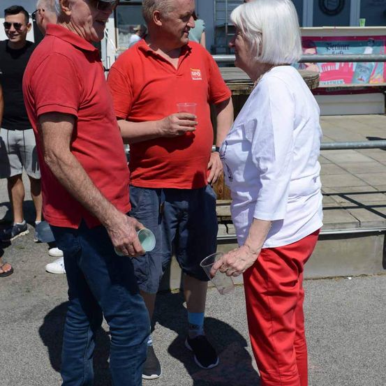 Zwei Männer und eine Frau stehen in einem Außenbereich. Die Frau hält einen Becher. Hinter ihnen stehen andere Menschen. Der Mann auf der linken Seite trägt eine Sonnenbrille und eine Uhr. Der Mann in der Mitte hält einen Becher. Die Frau trägt eine Halskette. Hinter ihnen befindet sich ein Holzzaun.