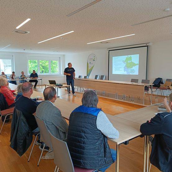 Ein Mann präsentiert eine Karte in einem Konferenzraum mit Teilnehmern, die um Tische herum sitzen.