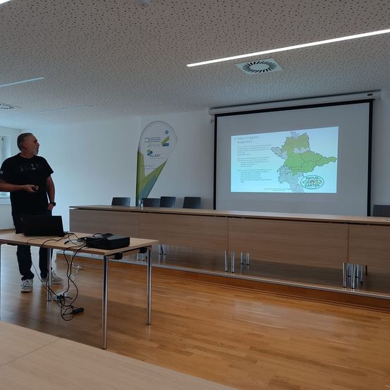 Ein Mann steht vor einem Holztisch mit Laptop und präsentiert eine Karte auf einer Projektionsleinwand in einem Konferenzraum.