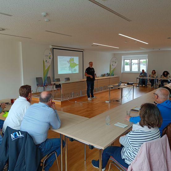 Eine Gruppe von Menschen sitzt in Stühlen um einen Konferenztisch herum und hört einem Mann in einem schwarzen T-Shirt zu, der neben einer Projektionsleinwand steht.