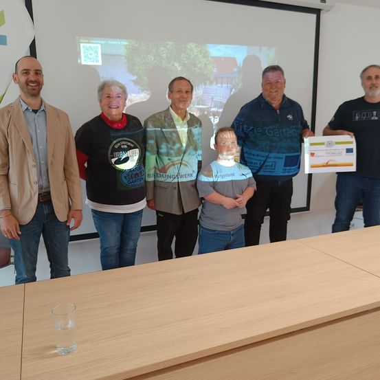 Eine Gruppe von sechs Personen steht in einem Raum mit einer Projektionsleinwand dahinter. Ein Junge hält ein Zertifikat und ein Mann steht neben ihm.