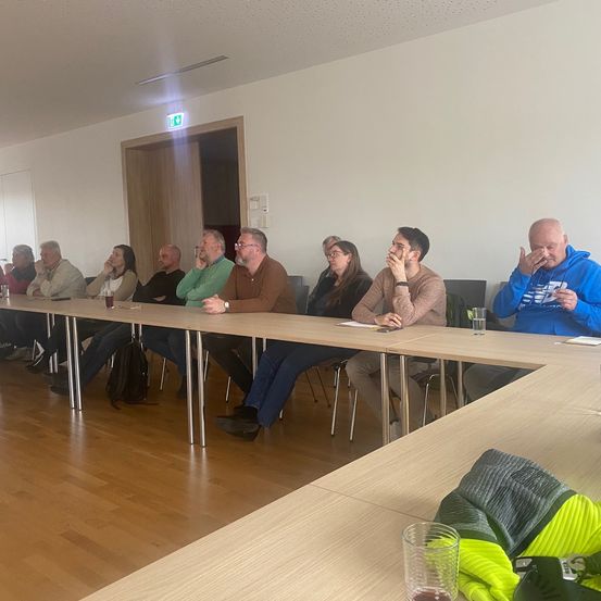 Eine Gruppe von Menschen sitzt in einem Raum, möglicherweise in einer Besprechung. Sie haben die Hände im Gesicht, einige trinken. Der Raum hat einen Holzboden und weiße Wände.