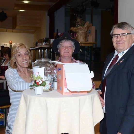 Drei Personen stehen an einem Tisch mit einem Blumenarrangement, einem Glasgefäß und einer Schachtel. Eine Frau in einem gestreiften Shirt lächelt in die Kamera. Ein Mann in Anzug und Krawatte schaut auf die Schachtel. Eine weitere Person steht hinter ihnen mit einem lächelnden Gesicht.