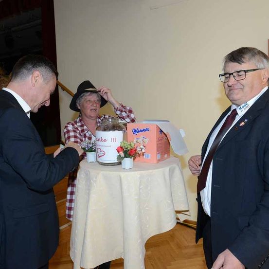 Drei Personen stehen um einen Tisch mit einem Glas und Blumen. Der Mann auf der linken Seite hält ein Stück Papier und der Mann auf der rechten Seite lächelt.
