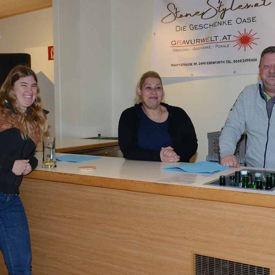 Drei Personen stehen lächelnd hinter einem Bartresen und posieren für ein Foto. Eine Frau auf der linken Seite hält ein Glas. Über ihnen befindet sich ein Banner mit dem Text 'Stone Stylesat'.