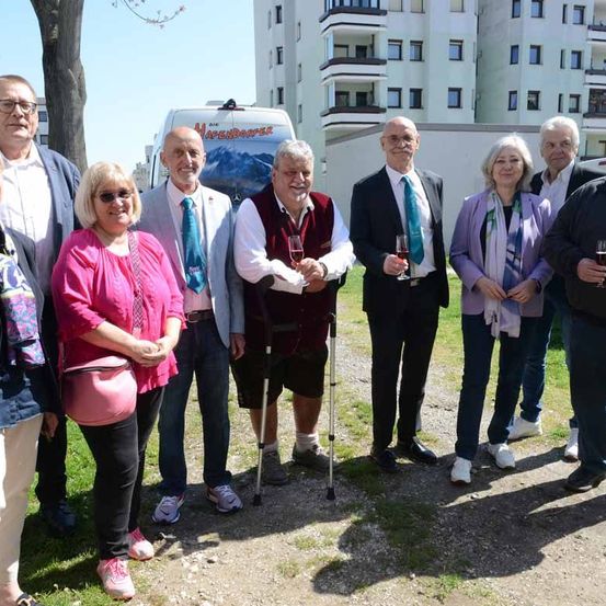 Eine Gruppe von Menschen steht vor einem weißen Gebäude, einige halten Weingläser, ein Mann benutzt einen Stock und ein Van ist dahinter geparkt.