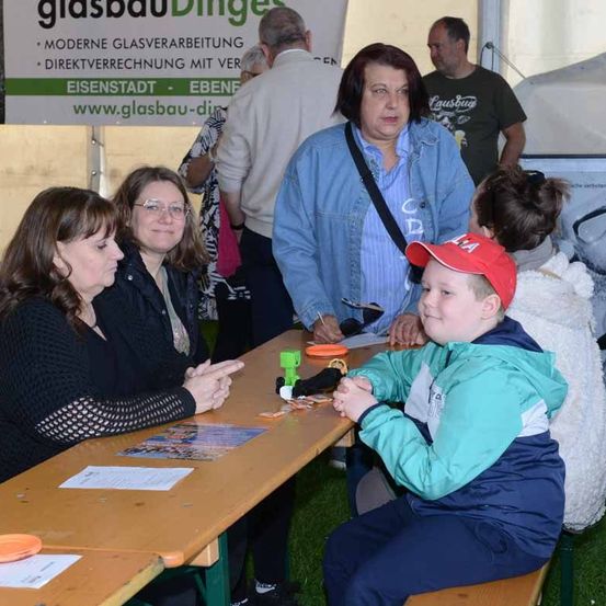 Eine Gruppe von Menschen versammelt sich an einem Tisch bei einer Veranstaltung im Freien, darunter eine erwachsene Frau mit Brille, ein Junge in einer roten Mütze und eine Frau in einer Jeansjacke.