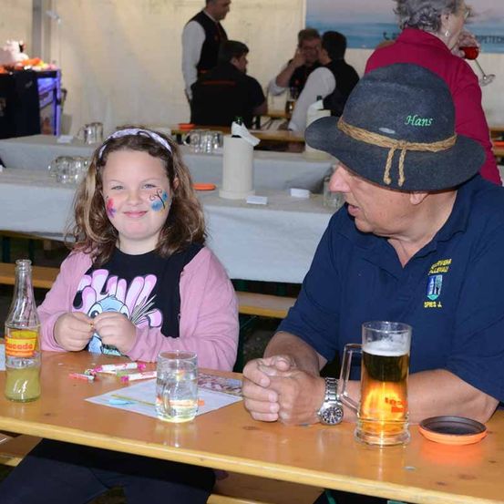 Ein Mädchen mit Gesichtsbemalung sitzt an einem Tisch mit einem Mann, der einen Hut mit dem Namen Hans trägt.