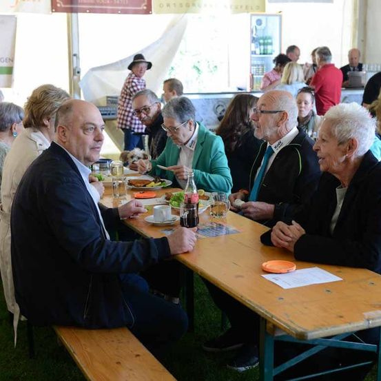 Eine Gruppe älterer Personen, darunter zwei ältere Frauen, sitzt an einem Tisch in einer Außenumgebung und genießt eine Mahlzeit.