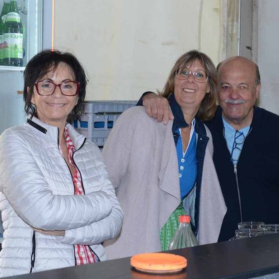 Drei Erwachsene, zwei Frauen und ein Mann, lächeln und posieren für ein Foto vor einem Kühlschrank mit Flaschen und einer Theke mit Gläsern und einem Teller.