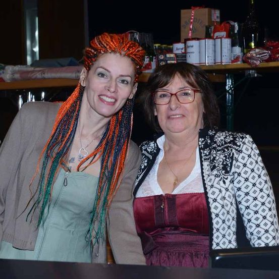 Zwei Frauen stehen gemeinsam und lächeln für ein Foto. Eine Frau hat bunte Zöpfe und eine Halskette. Die andere Frau trägt eine Brille und ein gemustertes Oberteil. Dahinter befindet sich ein Regal mit verschiedenen Gegenständen.