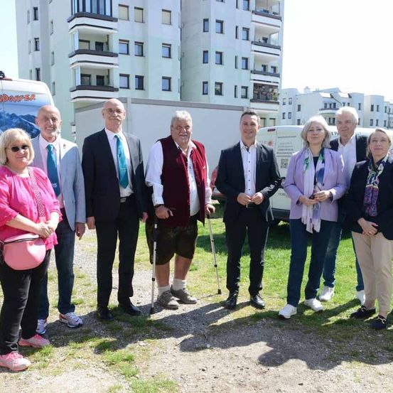 Eine Gruppe von Menschen in formeller und lässiger Kleidung steht auf einem Schotterplatz vor einem modernen Gebäude.