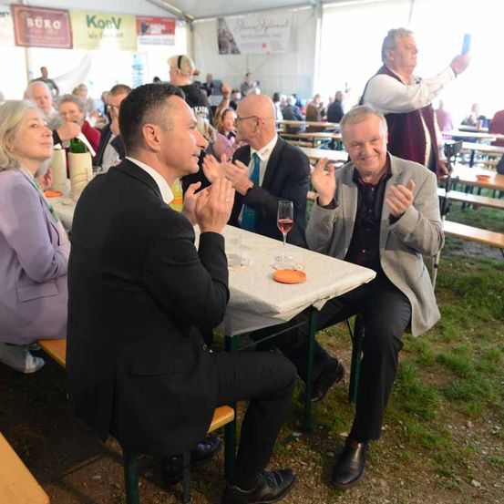 Zwei Männer sitzen auf einer Bank und klatschen in die Hände. Eine Frau sitzt neben ihnen. Ein Mann macht ein Foto mit seinem Handy.