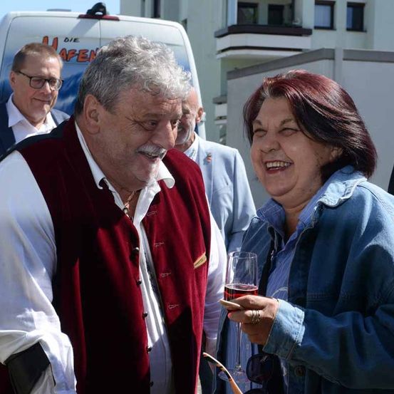 Ein Mann und eine Frau lächeln und lachen draußen, mit zwei anderen Männern im Hintergrund. Die Frau hält ein Weinglas, und der Mann trägt eine rote Weste.