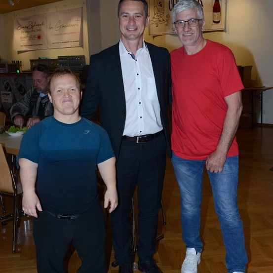 Drei Männer stehen zusammen in einem Raum. Der Mann in der Mitte trägt ein weißes Hemd und der Mann rechts trägt ein rotes Hemd. Hinter ihnen sitzt ein Mann am Tisch.