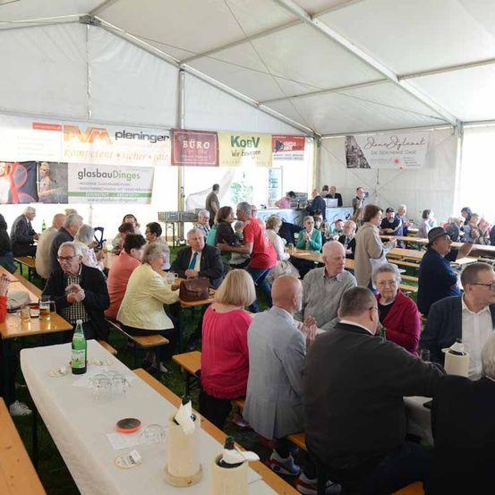 Viele ältere Menschen sitzen in einem Zeltrestaurant und unterhalten sich. Auf den Tischen stehen mehrere Flaschen und Gläser, außerdem hängen mehrere Banner an den Wänden.
