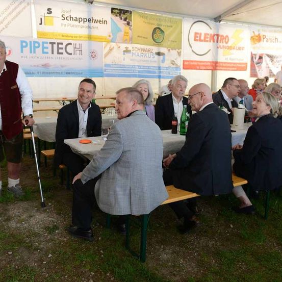 Eine Gruppe von Menschen sitzt unter einem Zelt an Tischen. Ein Mann benutzt einen Stock und ein anderer hält eine Flasche. Im Hintergrund sind verschiedene Banner sichtbar.